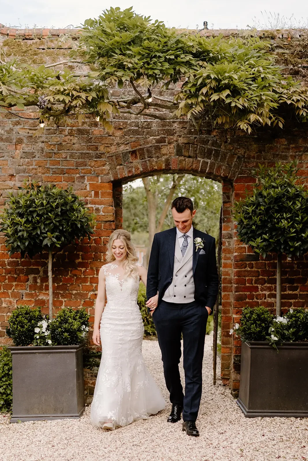A bride and groom walking hand in hand through a brick archway with greenery and potted plants on either side, celebrating their wedding day.