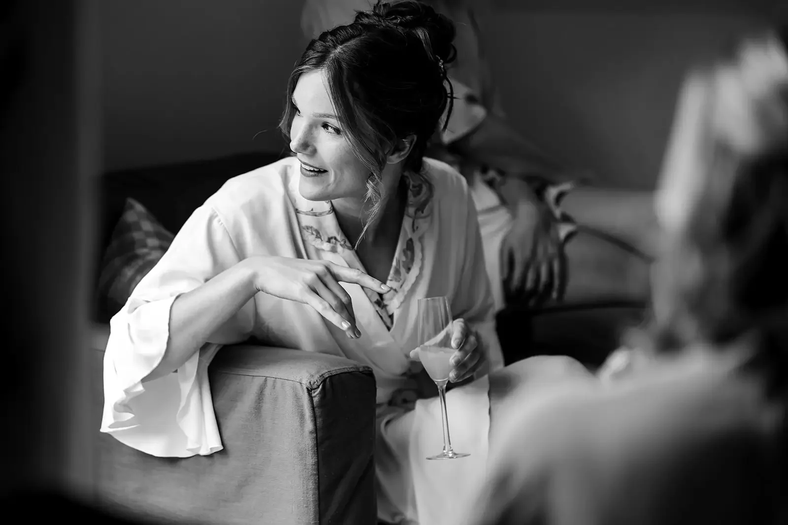 A woman smiling and holding a glass of champagne while sitting on a sofa, engaged in conversation with someone partially visible in the foreground, in a social setting.