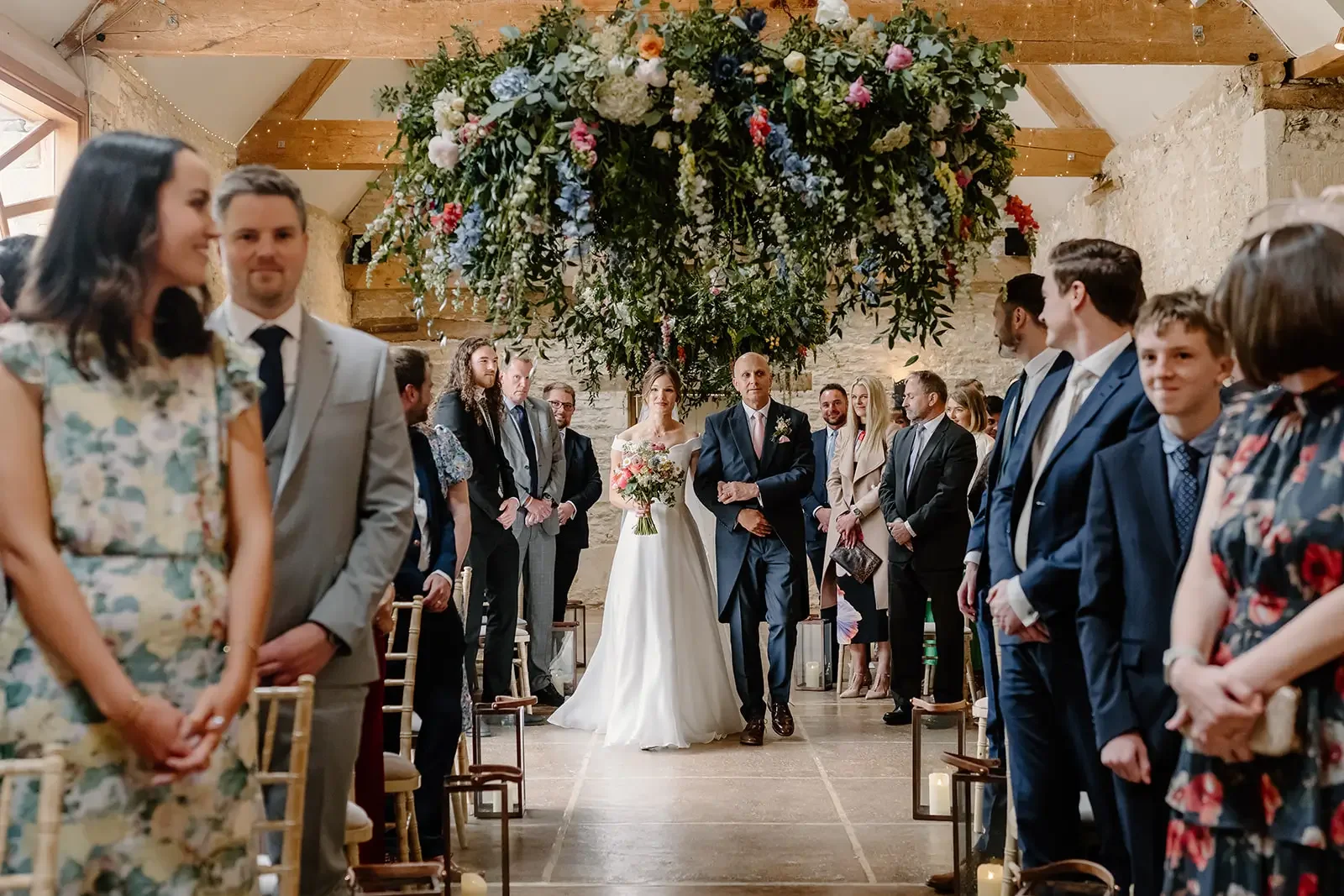Bride walking down the aisle with her father at a wedding ceremony in a rustic indoor venue decorated with a large floral arrangement overhead.