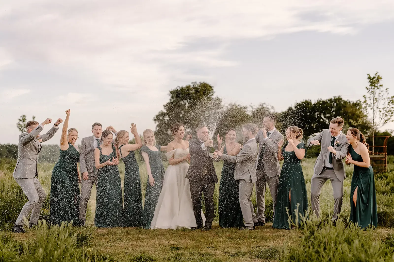Group of people celebrating outdoors, with some spraying champagne, during a wedding or similar event.