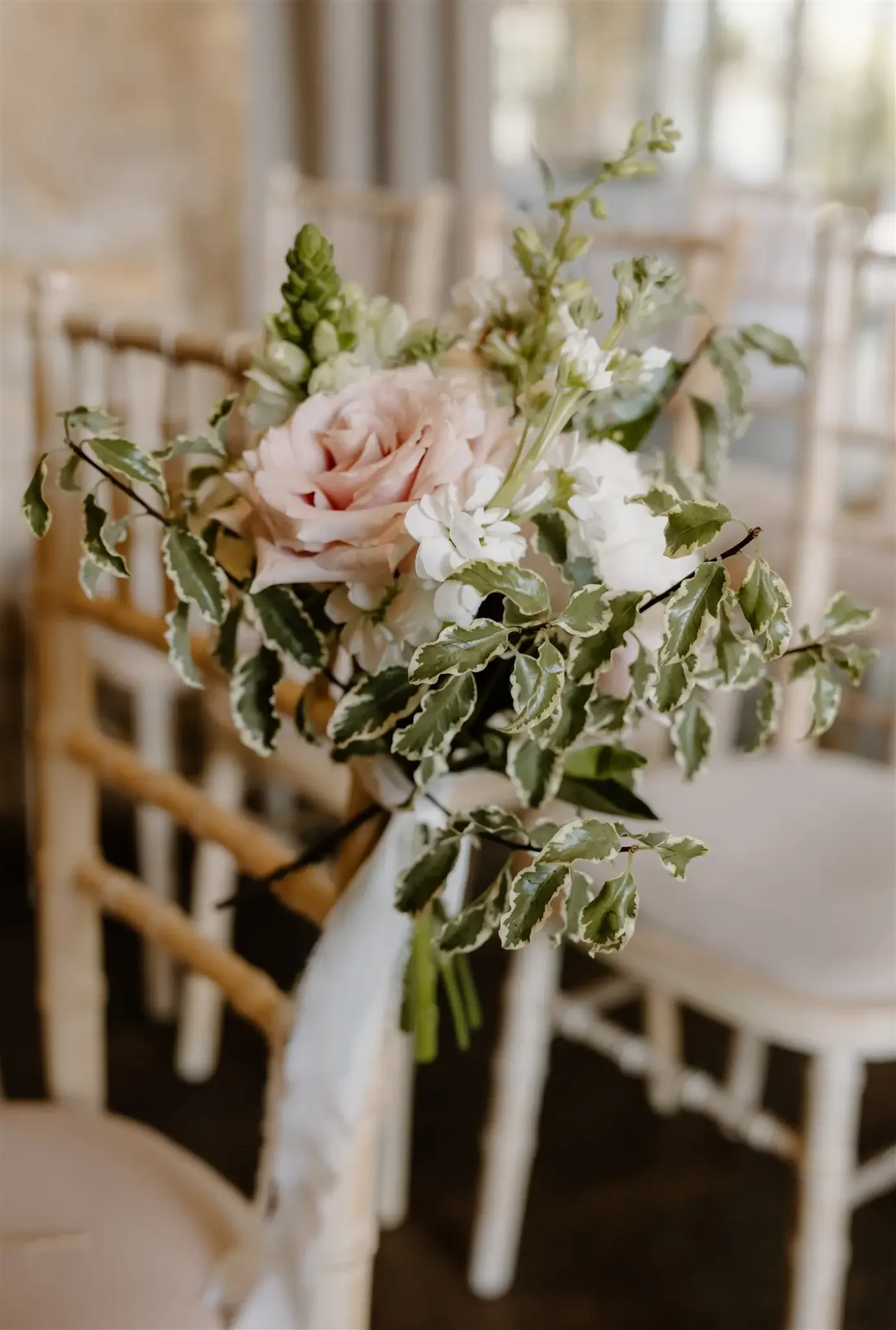 A bouquet of pink and white flowers with variegated green and white leaves, attached to a chair at a decorated venue.