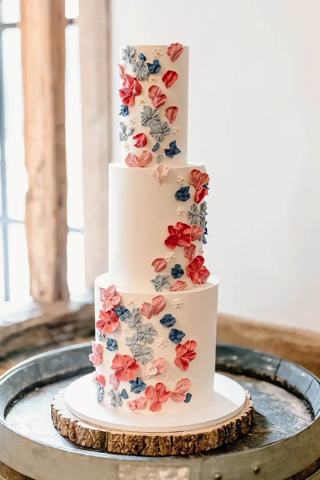 A tall white multi-tiered wedding cake decorated with pink, blue, and gray sugar flowers, placed on a wooden slab on a metal tray.