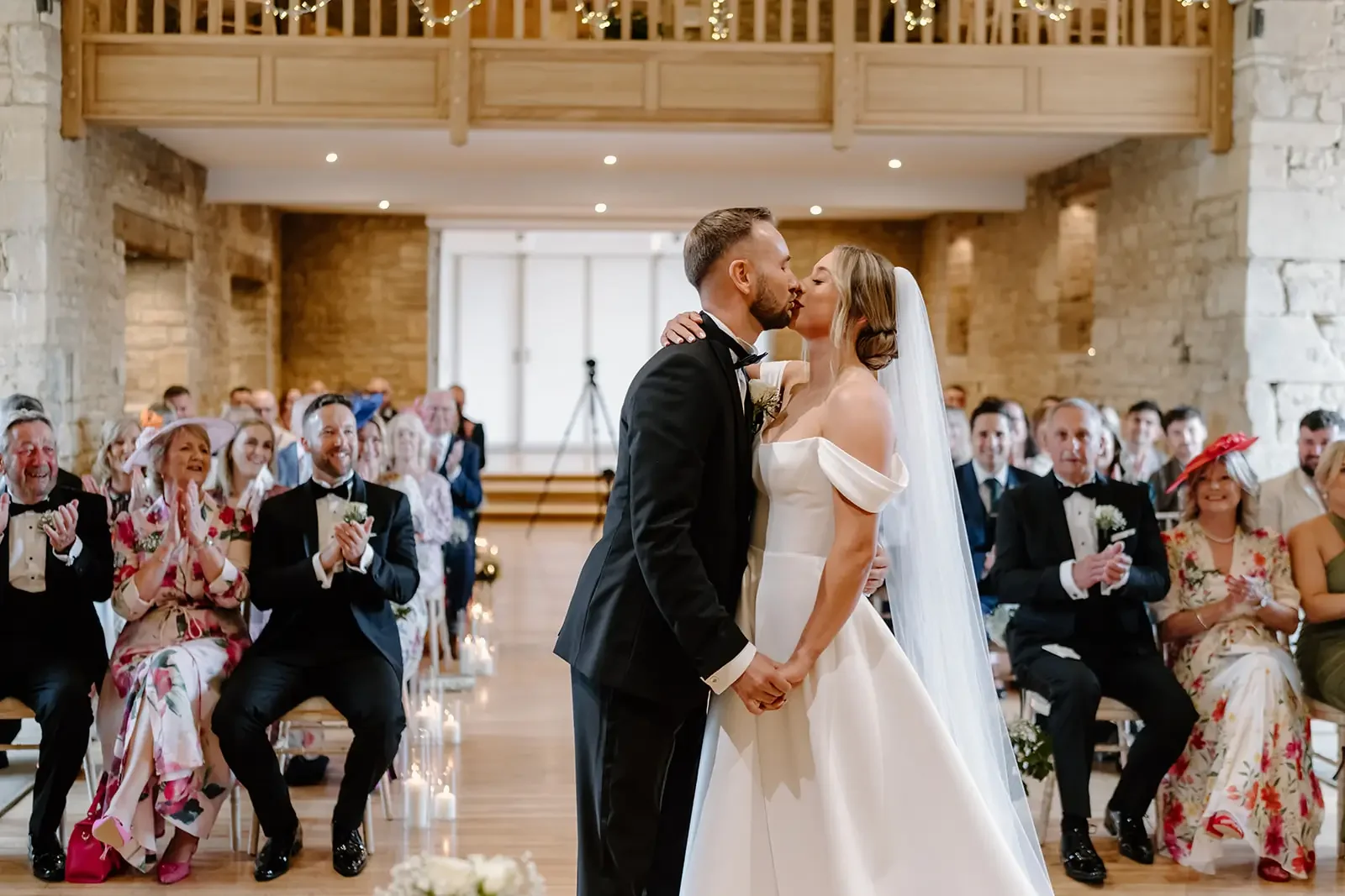 A bride and groom kiss during their wedding ceremony in a rustic venue with exposed brick walls. Guests seated in the background are clapping and smiling.