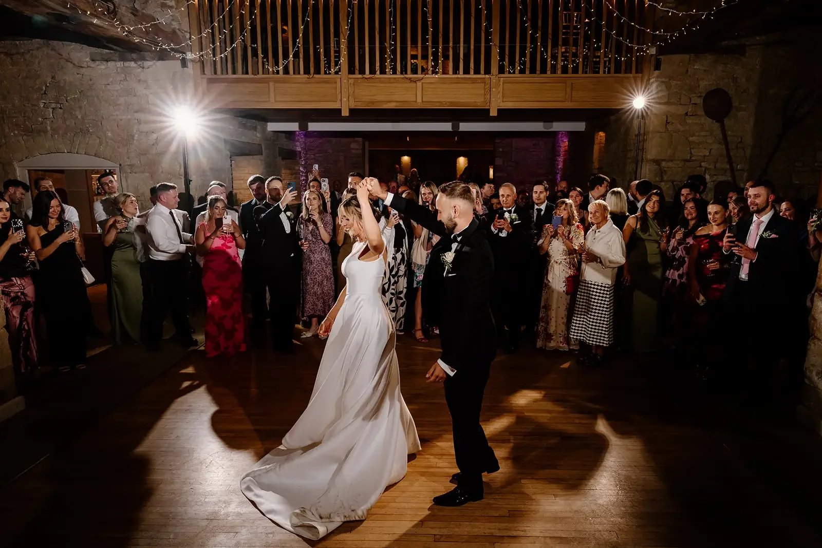 A bride and groom are dancing at their wedding reception, surrounded by guests taking photos and watching in a rustic venue with wooden beams and stone walls.