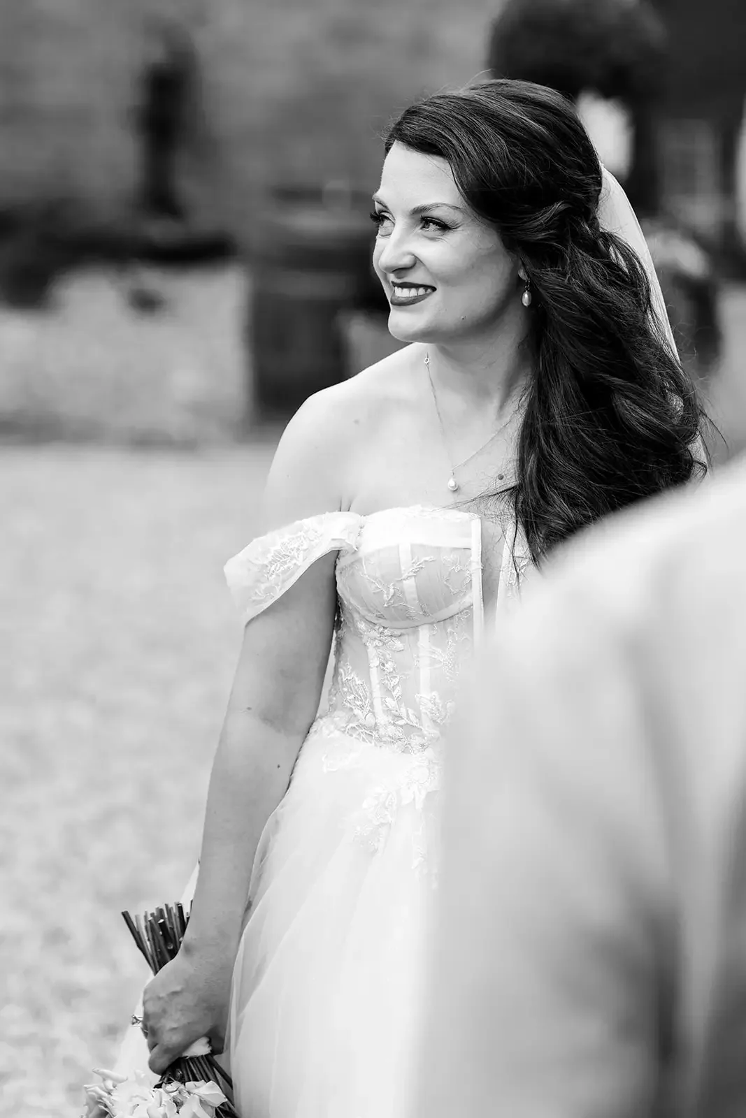 Black and white photo of a bride smiling, holding a bouquet of flowers, wearing an off-the-shoulder lace wedding dress with long dark hair styled in loose waves.