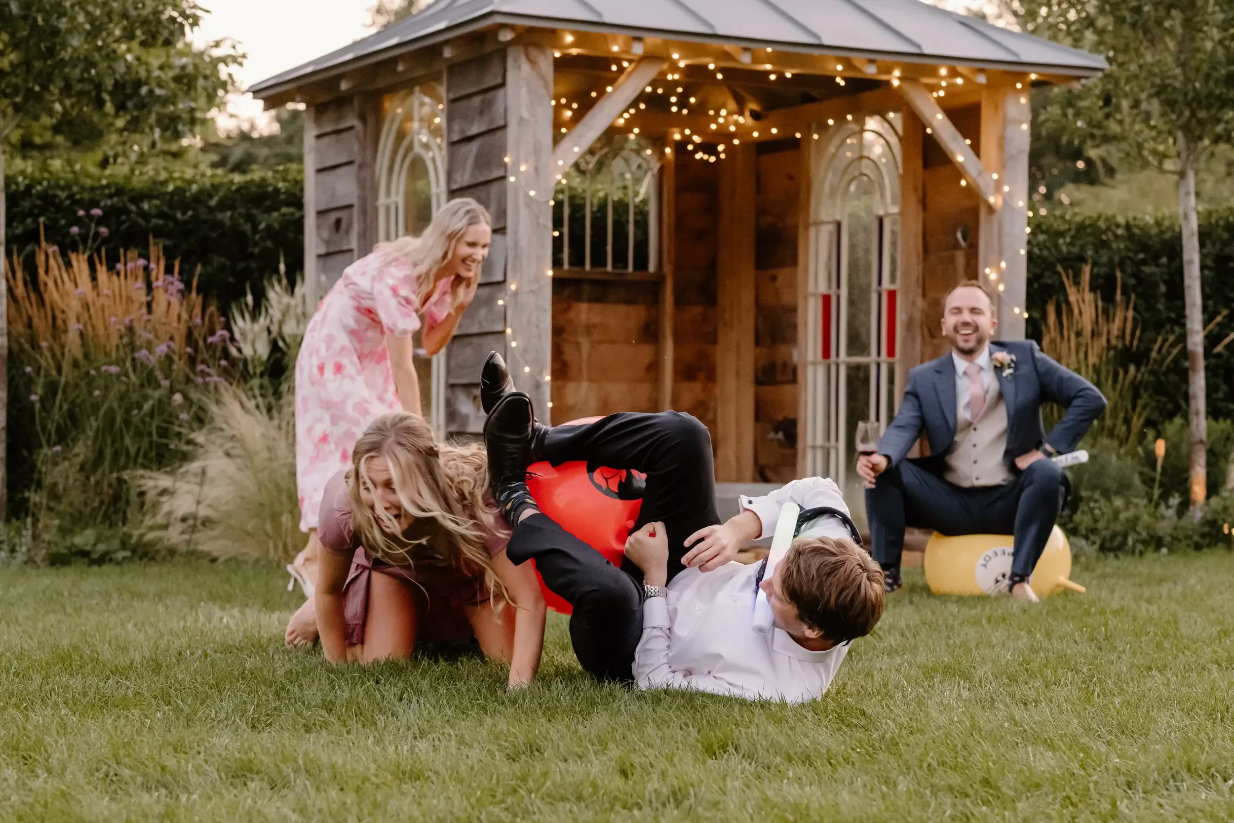 People playing on the grass outdoors during a celebration, with a small wooden shed decorated with string lights in the background. One person is lying on the ground while others are engaged around them, and a man in a suit is sitting on a yellow tea