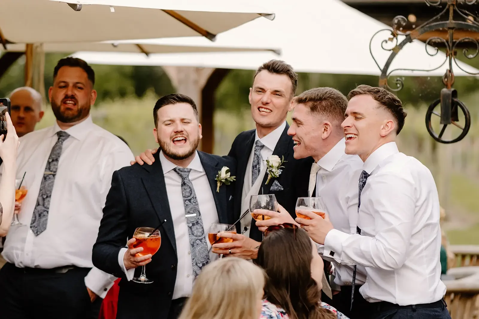 A group of seven men in formal suits celebrating and smiling, holding drinks, at an outdoor wedding or party with umbrellas and greenery in the background.