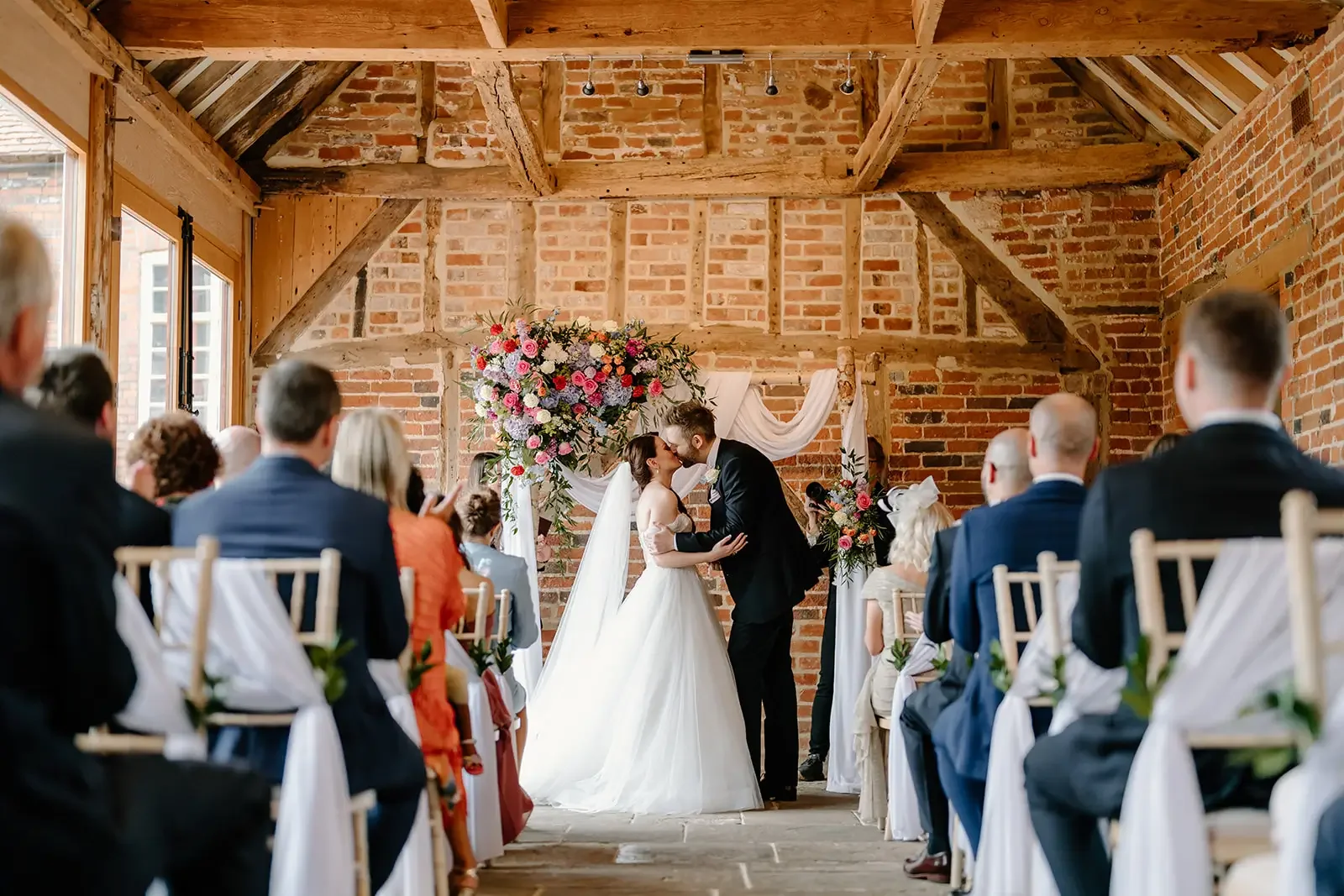 A bride and groom sharing a kiss at their wedding ceremony inside a rustic venue with exposed brick walls and wooden beams, surrounded by seated guests.