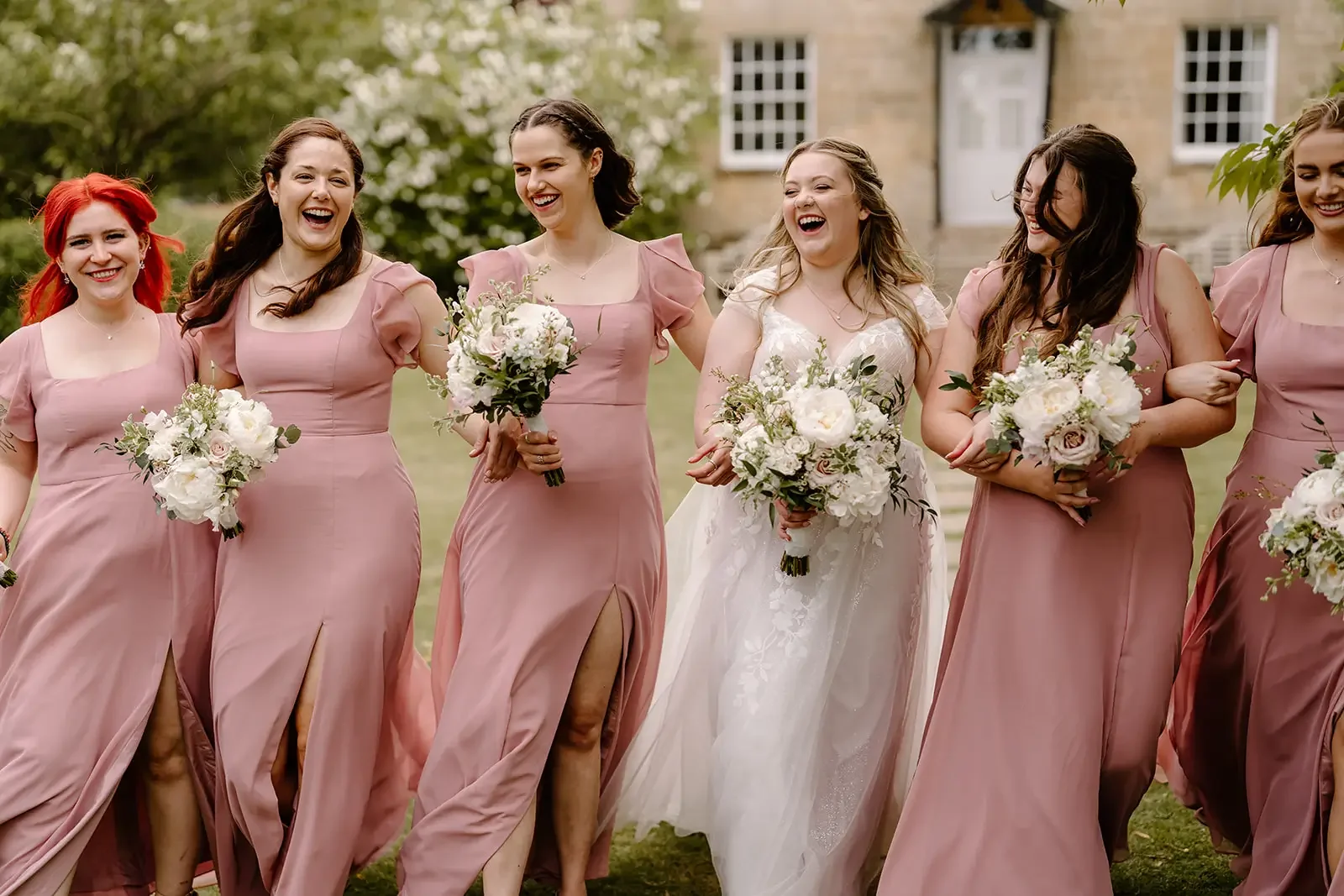 A group of six women at a wedding, dressed in pink bridesmaid dresses and a white wedding gown, holding bouquets, walking arm in arm outdoors in front of a house with a garden.