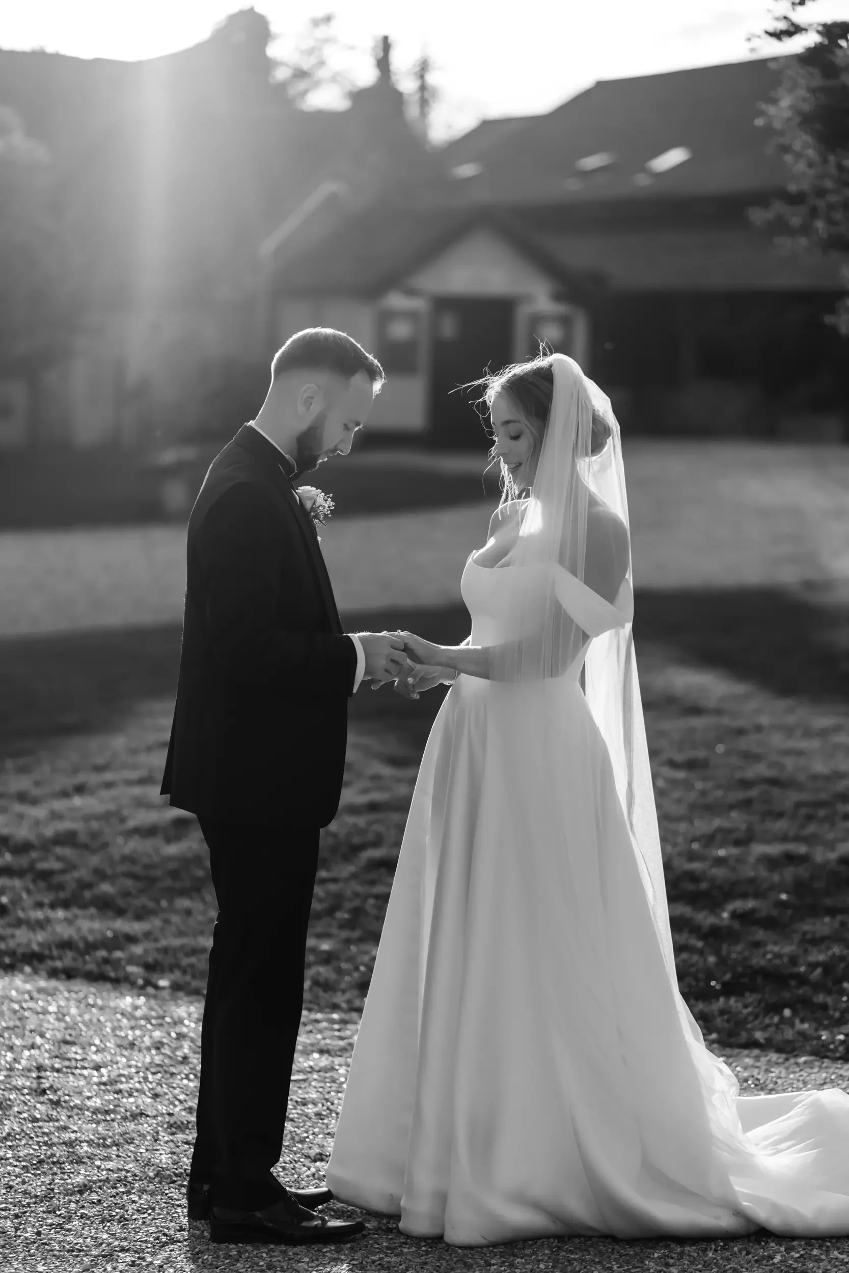 A black and white photo of a bride and groom standing outdoors, holding hands and looking down at each other in front of a house, with sunlight shining behind them.