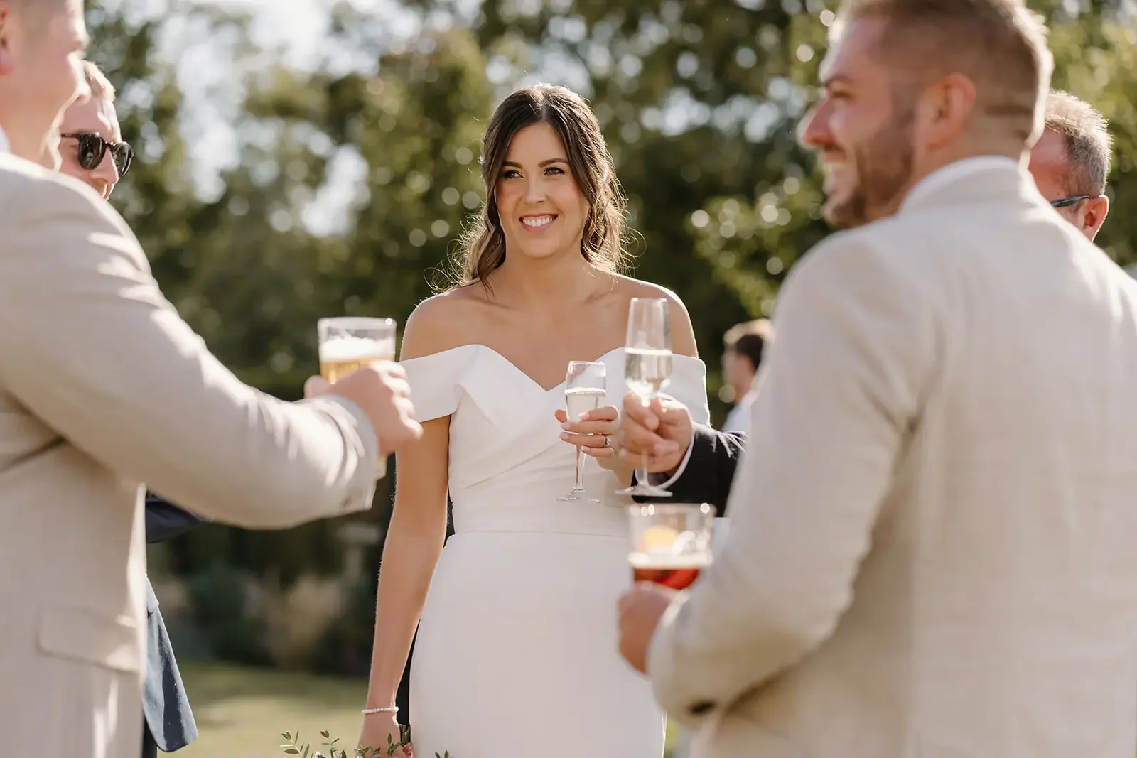 A woman in a white dress holding a glass of champagne, smiling, while surrounded by men in light-colored suits holding drinks at an outdoor wedding reception.