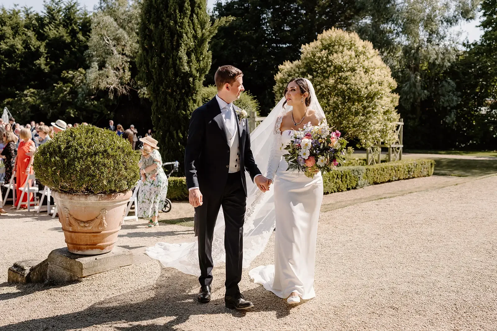 Bride and groom holding hands and walking outdoors during their wedding ceremony, surrounded by guests and lush greenery.