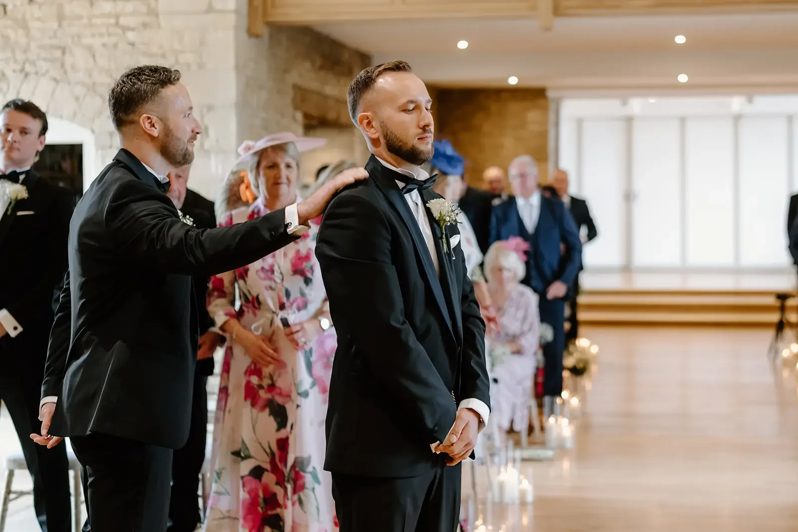 Groom in a black tuxedo with a white boutonniere standing with eyes closed as another man in a tuxedo touches his shoulder during a wedding ceremony in a bright, decorated venue.