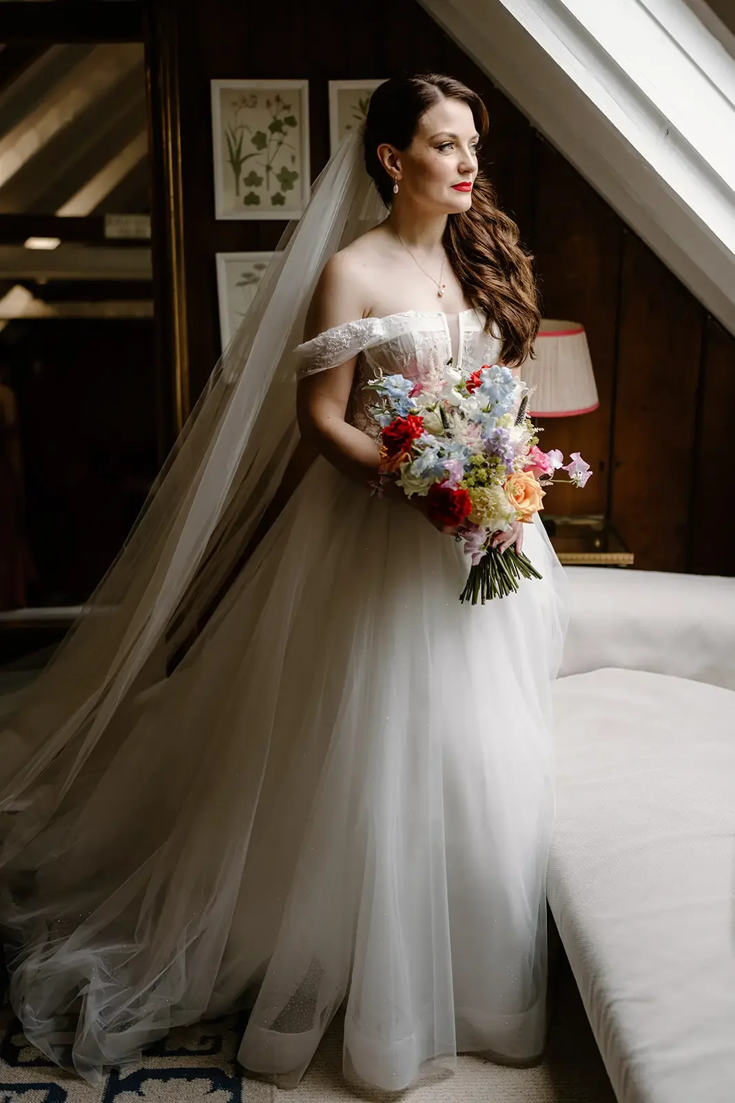 A bride with long, wavy brown hair in an off-the-shoulder wedding dress holding a bouquet of colorful flowers, standing near a window with a sloped ceiling and dark wood paneling.