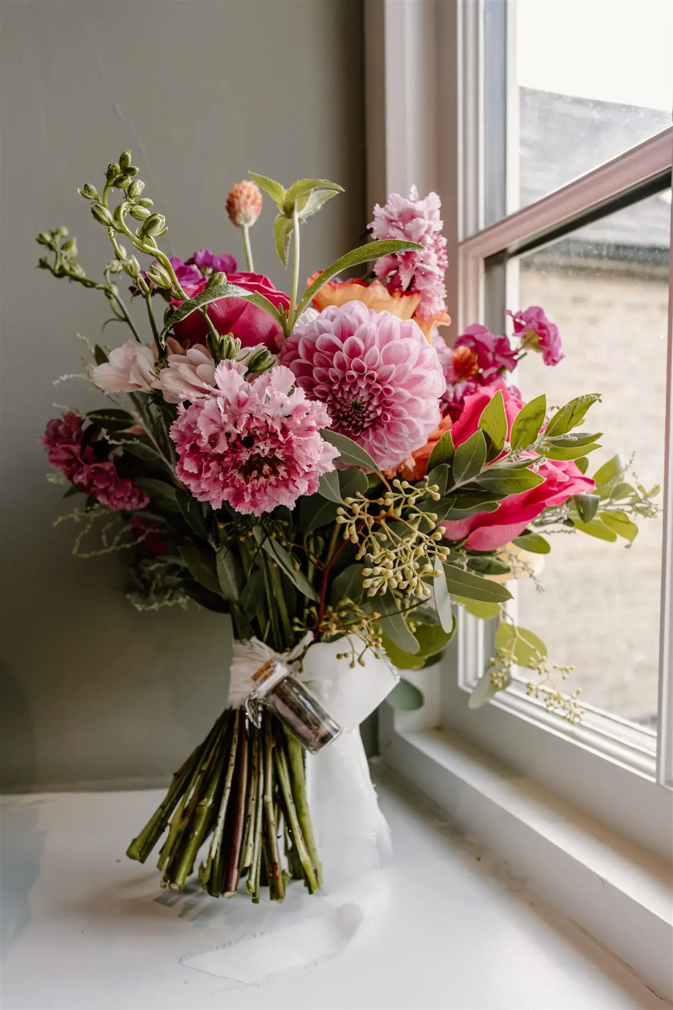 A bouquet of pink flowers in a white vase placed on a windowsill.