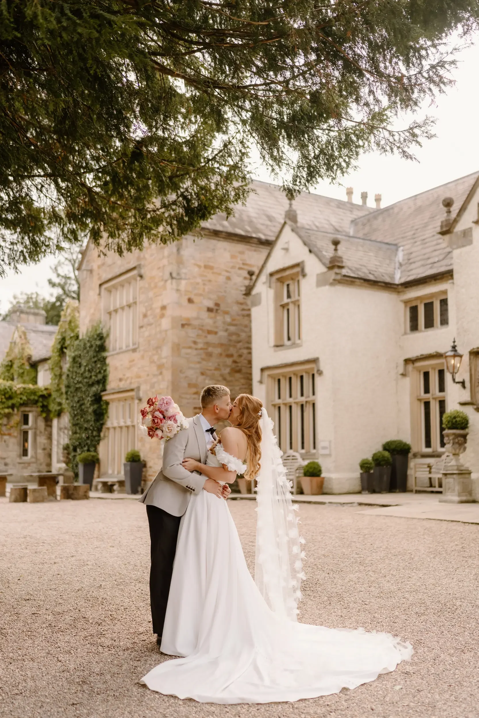 A bride and groom sharing a kiss outside a historic stone building on their wedding day, the bride holding a bouquet of pink and white flowers.