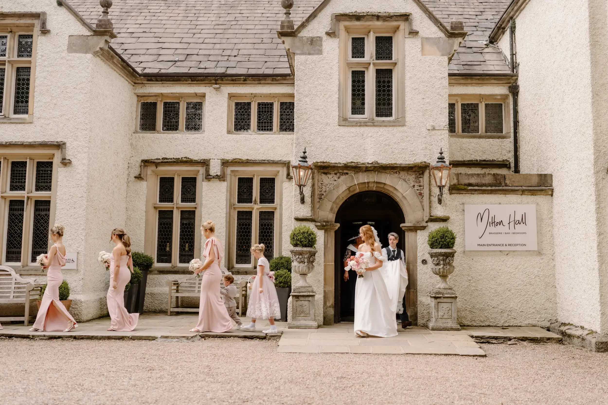 A bride in a white wedding dress leading a procession of bridesmaids in pink dresses and young flower girls outside a historic stone building with a sign that reads "Mitton Hall."