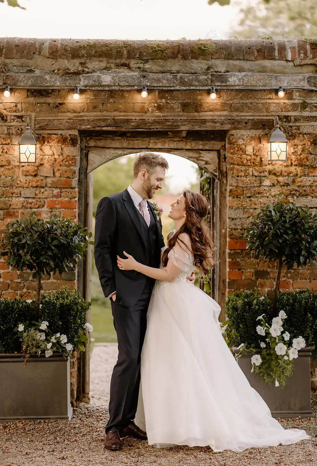 A bride and groom embracing each other at their wedding outdoors, standing in front of a brick archway decorated with string lights and lanterns, surrounded by potted plants and flowers.