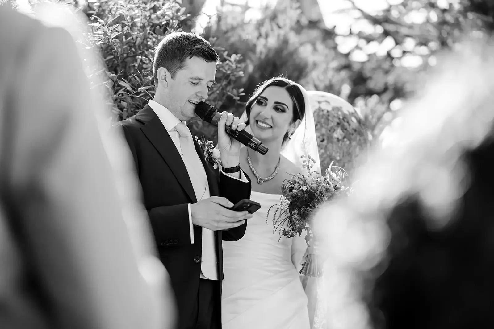 A black-and-white photo of a wedding ceremony outdoors, with a groom reading vows into a microphone while holding a phone, and a bride smiling at him holding a bouquet of flowers.