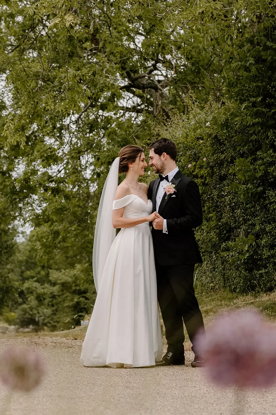 A bride and groom stand close together outdoors, holding hands, with faces touching and smiling. The bride is in a white off-the-shoulder wedding gown with a veil, and the groom is in a black tuxedo with a bow tie. They are surrounded by green foliag