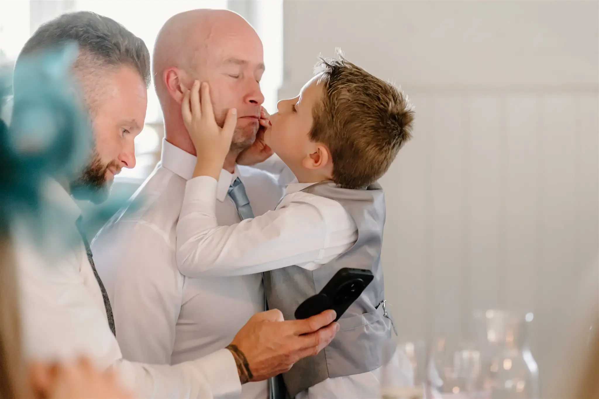 Young boy giving an older man a kiss on the lips, with a third man smiling in the background. The older man is holding a phone, and all three are dressed in formal attire, in a bright, indoor setting.