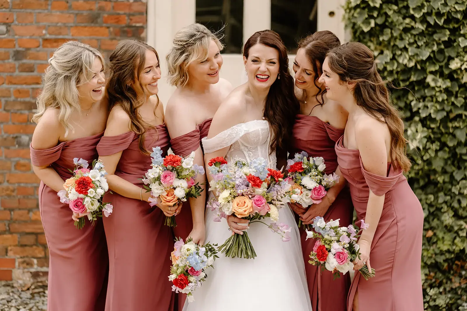 A bride in a white wedding dress surrounded by six bridesmaids in mauve dresses, all holding colorful bouquets, standing outdoors with a brick wall and greenery in the background.