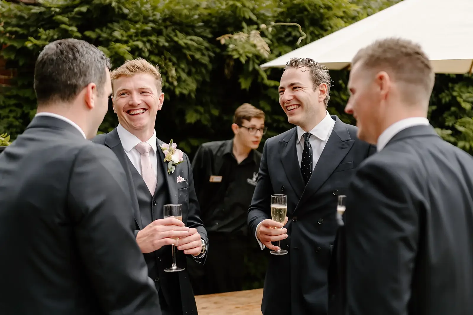 Four men in suits conversing and smiling at an outdoor event, holding glasses of champagne, with a woman in black in the background and green foliage and a white umbrella.