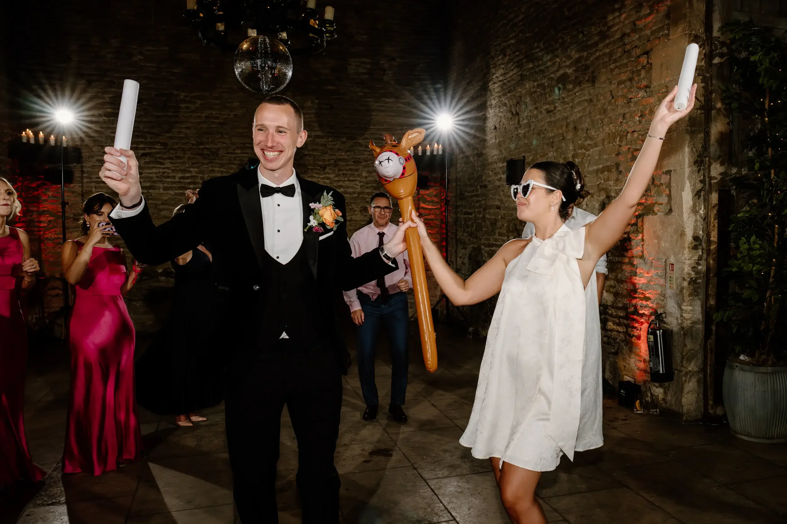 Groom and bride dancing at wedding reception, holding inflatable microphone and horse toy, in a rustic venue with other guests in the background.