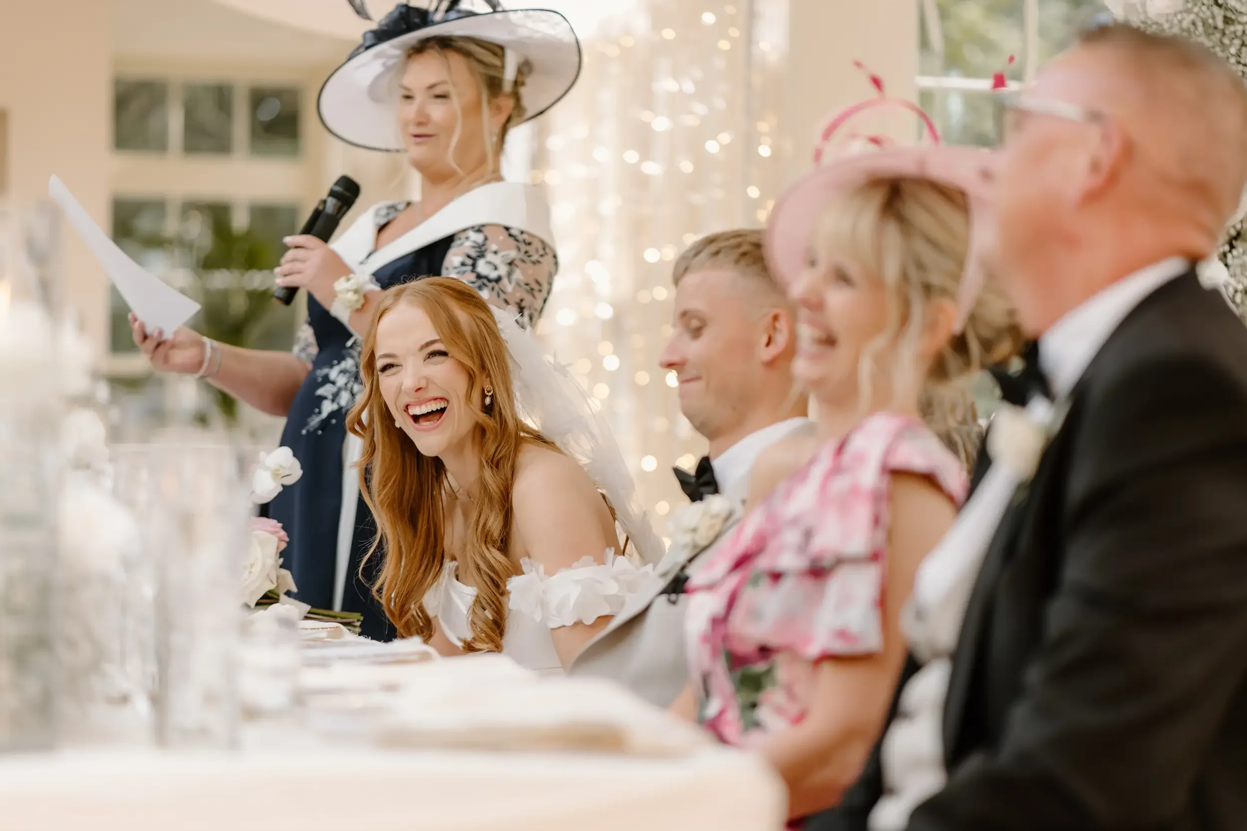 A group of five people at a wedding reception, seated at a table, smiling and enjoying the moment. The woman in the foreground has long red hair and is wearing a white dress, laughing. Other guests include a man in a tuxedo, a woman with a pink hat, 
