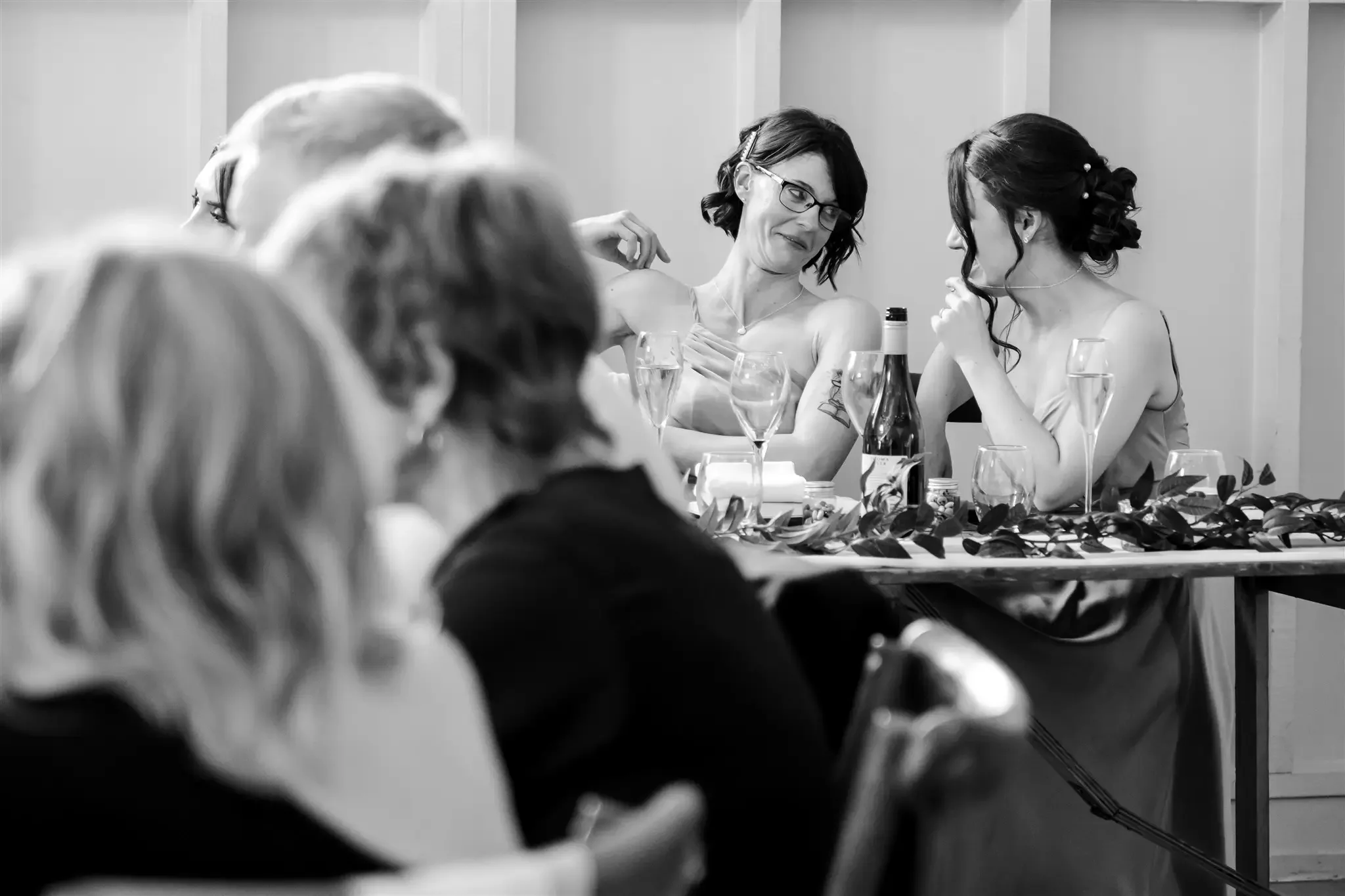 Black-and-white photo of a wedding reception with a bride and maid of honor sitting at a table decorated with flowers and glasses, engaged in conversation, with other guests in the foreground.
