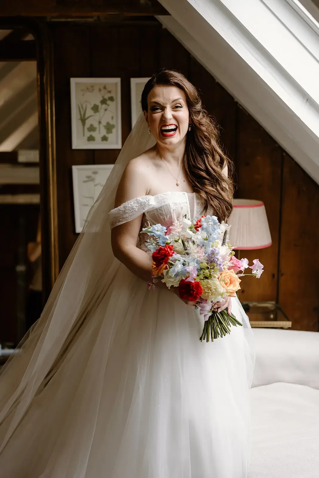 A bride in an off-the-shoulder wedding gown holding a colorful bouquet, laughing indoors near a large window with wooden walls and framed botanical prints behind her.