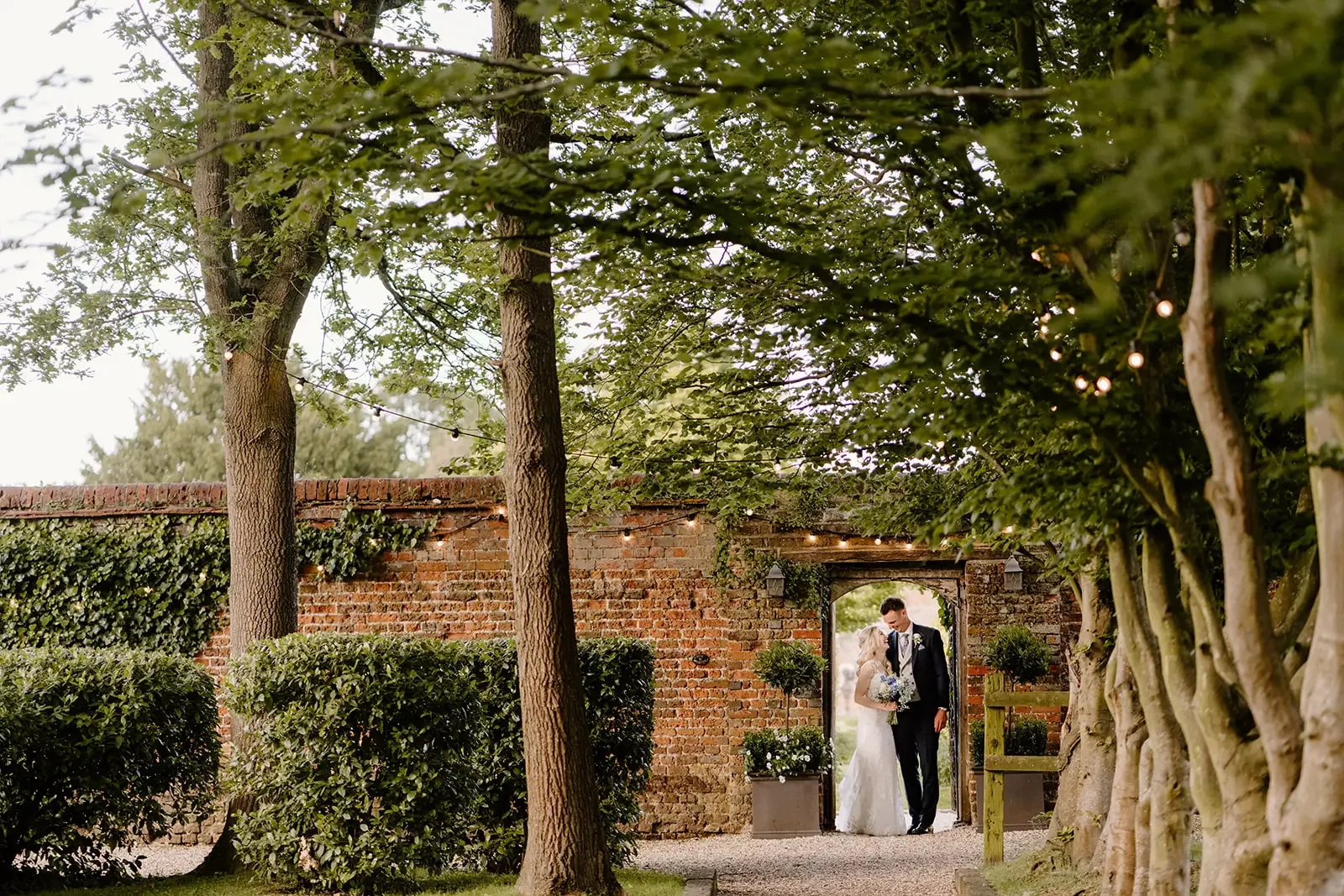 A bride and groom standing under an archway in a garden setting with trees and string lights overhead.