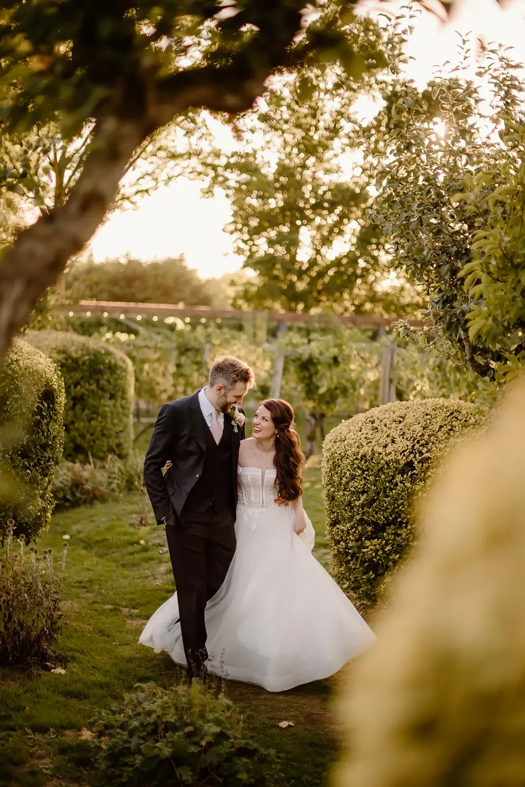 A newlywed couple walking together through a garden at sunset, smiling and looking at each other. The bride is in a white wedding dress and the groom is in a dark suit.
