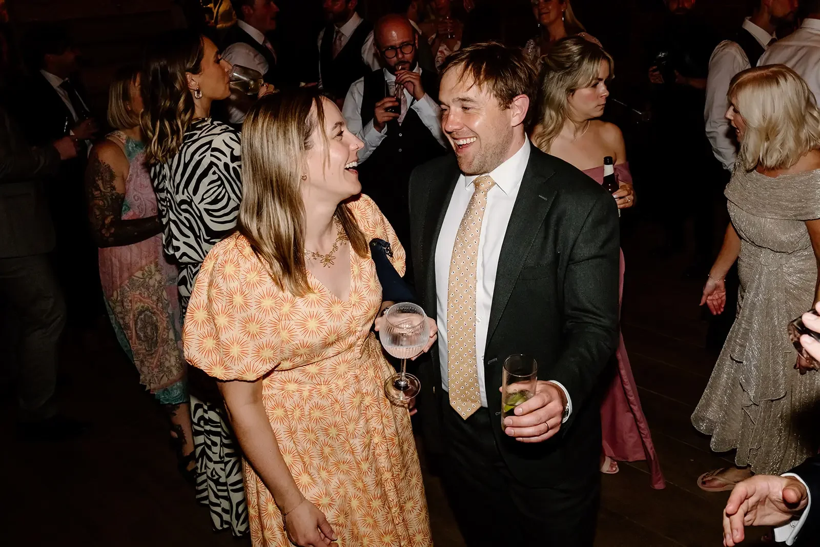 Man and woman smiling and looking at each other at a social event, holding drinks, surrounded by other people in formal attire.