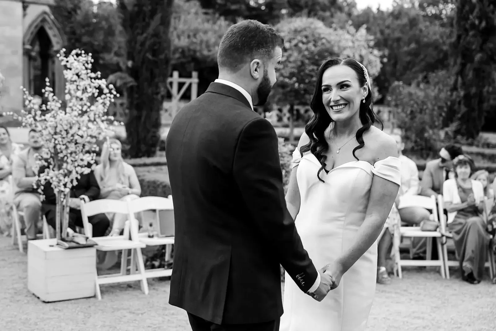 A bride and groom holding hands and exchanging vows at an outdoor wedding ceremony with guests seated in the background.