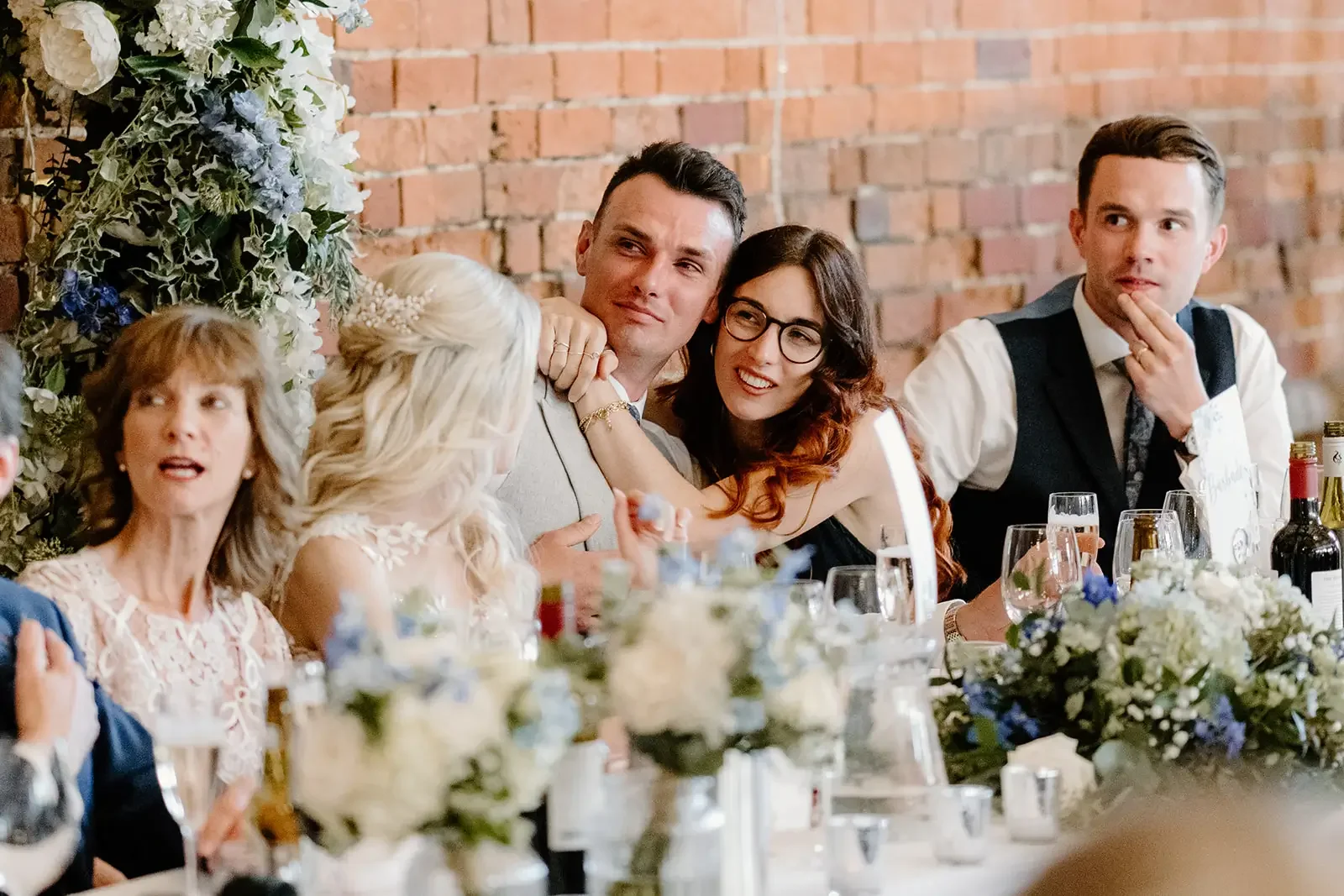 People sitting at a wedding reception, with a woman in a wedding dress, and guests seated at a table with flowers and drinks.