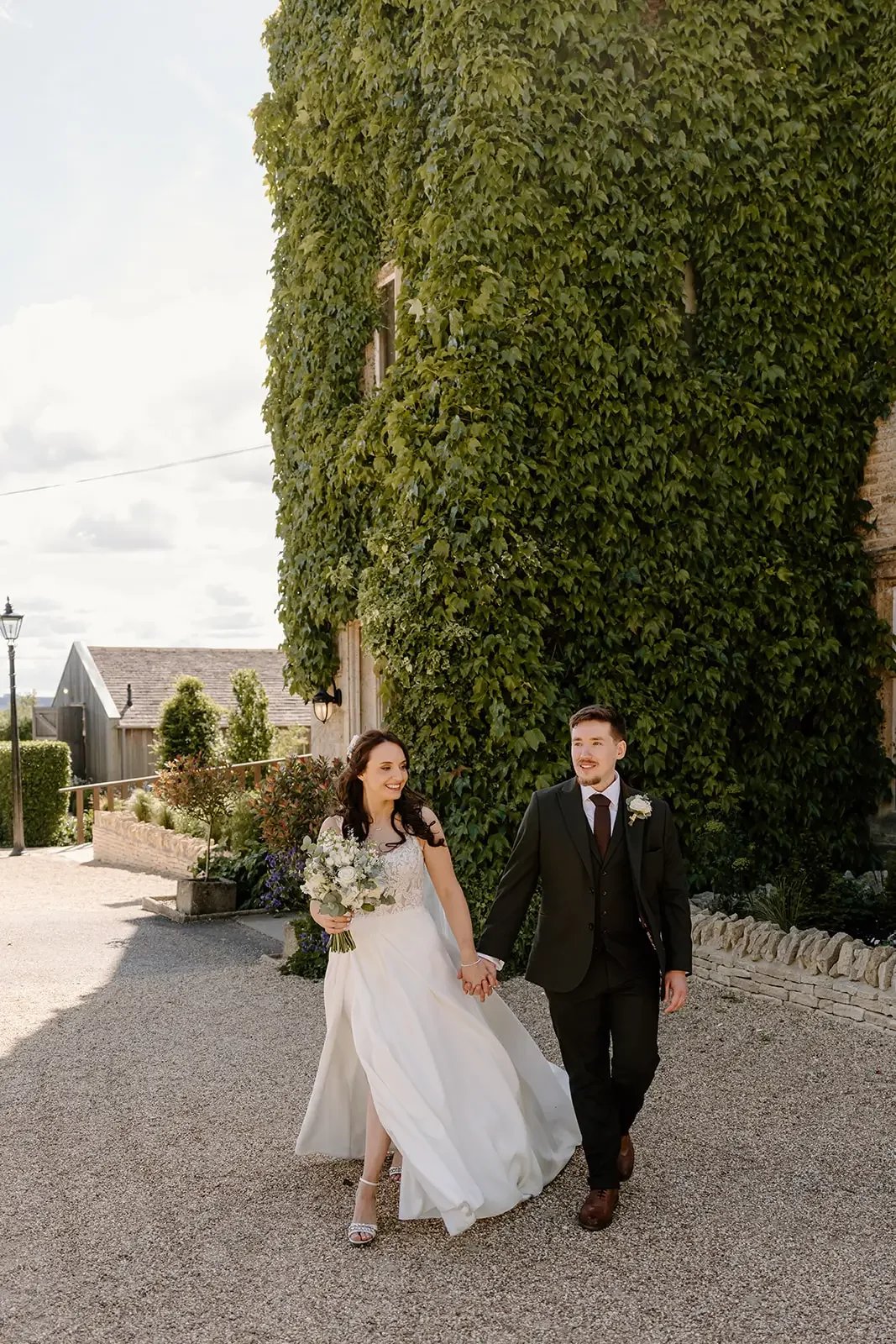 A bride and groom smiling and holding hands, walking outdoors on a sunny day, with a background of green ivy-covered building, garden plants, and a clear sky.