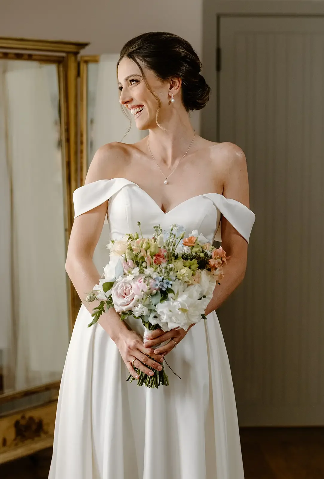 A woman in a white wedding dress holding a bouquet of flowers, smiling and looking to the side indoors.