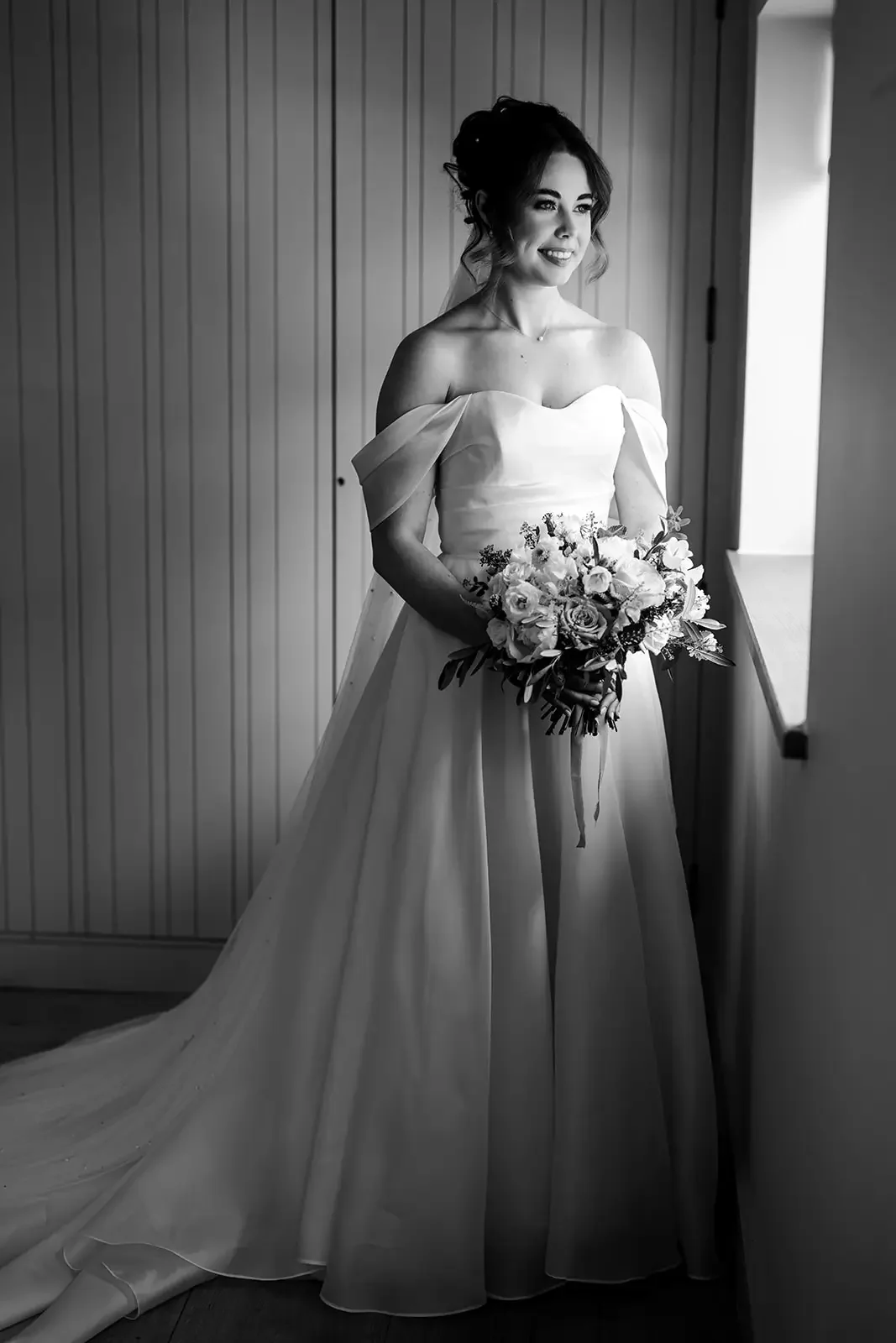 Black and white photo of a bride in an off-shoulder wedding gown holding a bouquet of flowers, standing near a window with wood-paneled walls.