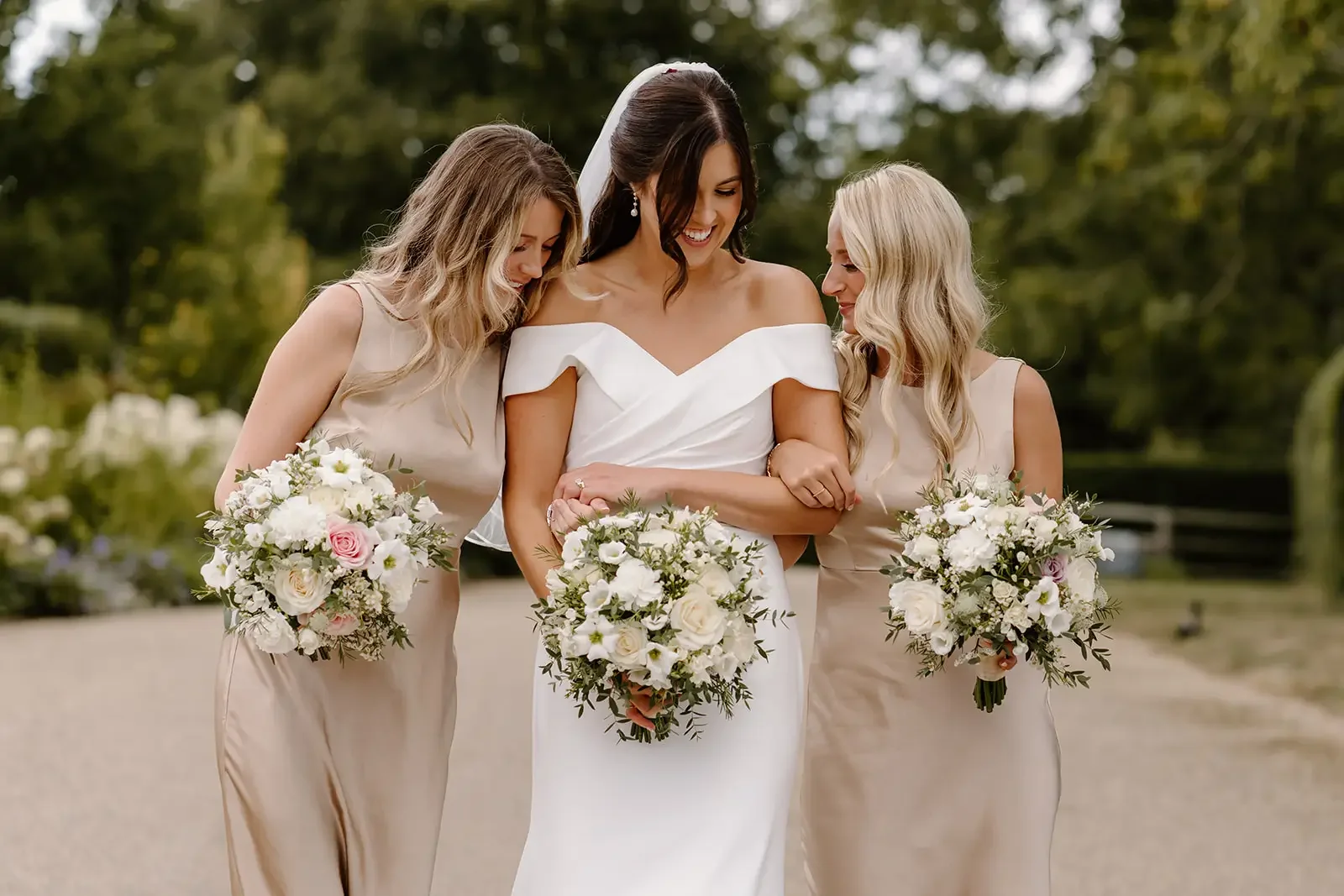 A bride in a white wedding dress walking with two bridesmaids in beige dresses on a pathway outdoors, all holding bouquets of white and pink flowers, surrounded by green trees.
