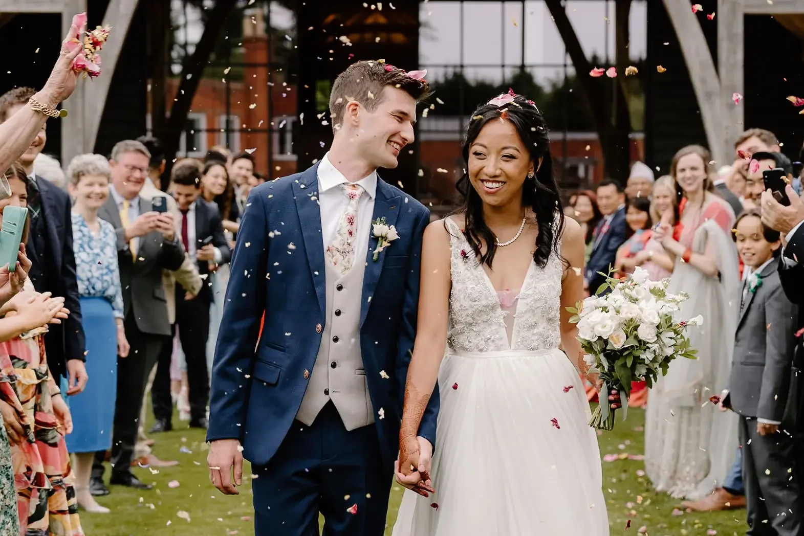 A bride and groom walking hand in hand at their wedding ceremony, surrounded by guests throwing flower petals and taking photos outdoors.