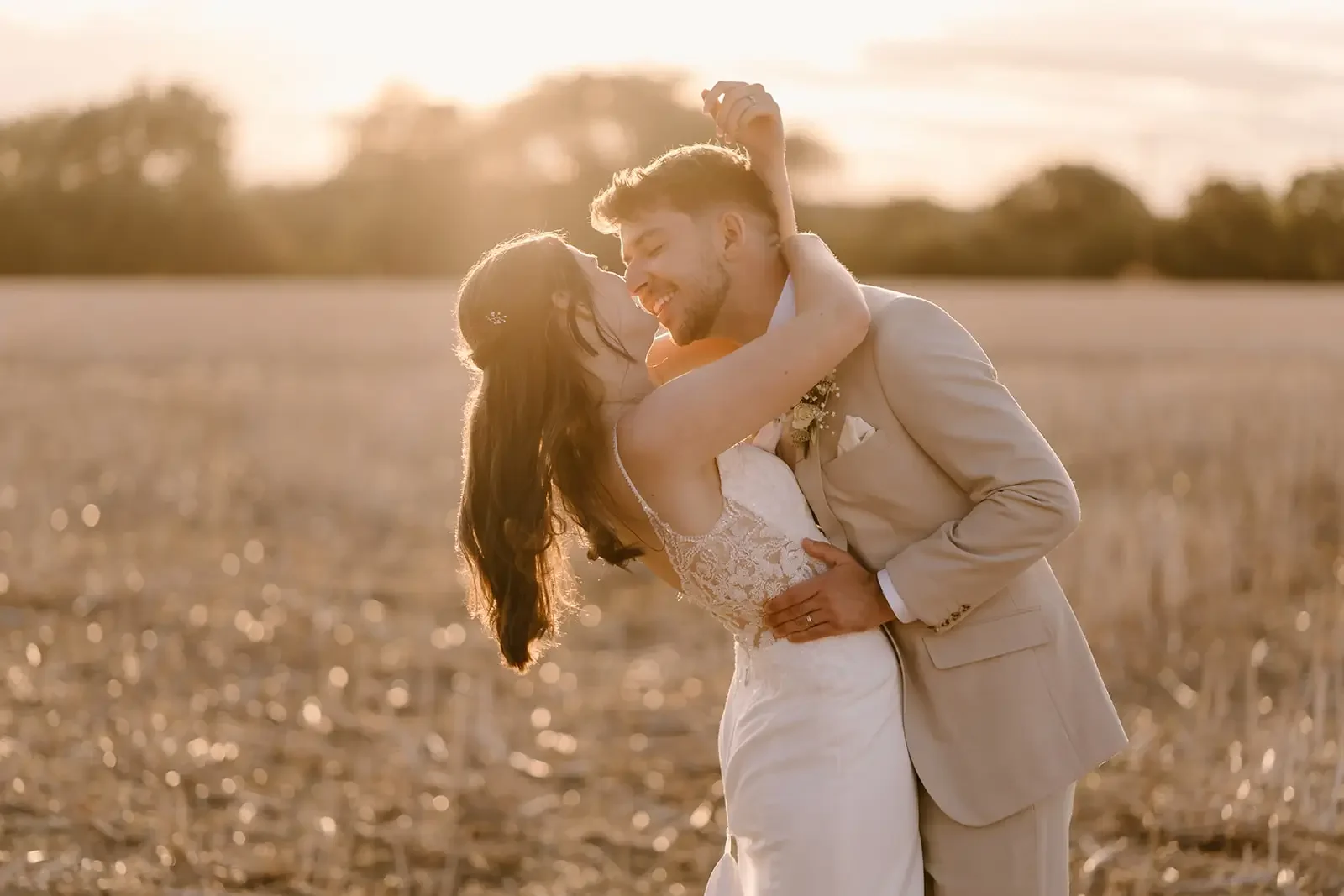 A newlywed couple in wedding attire sharing a dance in a field at sunset, embracing each other.
