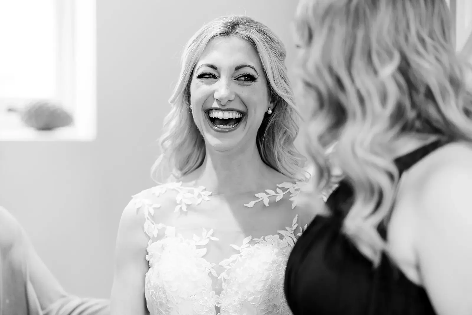 A woman in a wedding dress smiling and laughing while talking with another woman with wavy hair at a wedding or celebration.