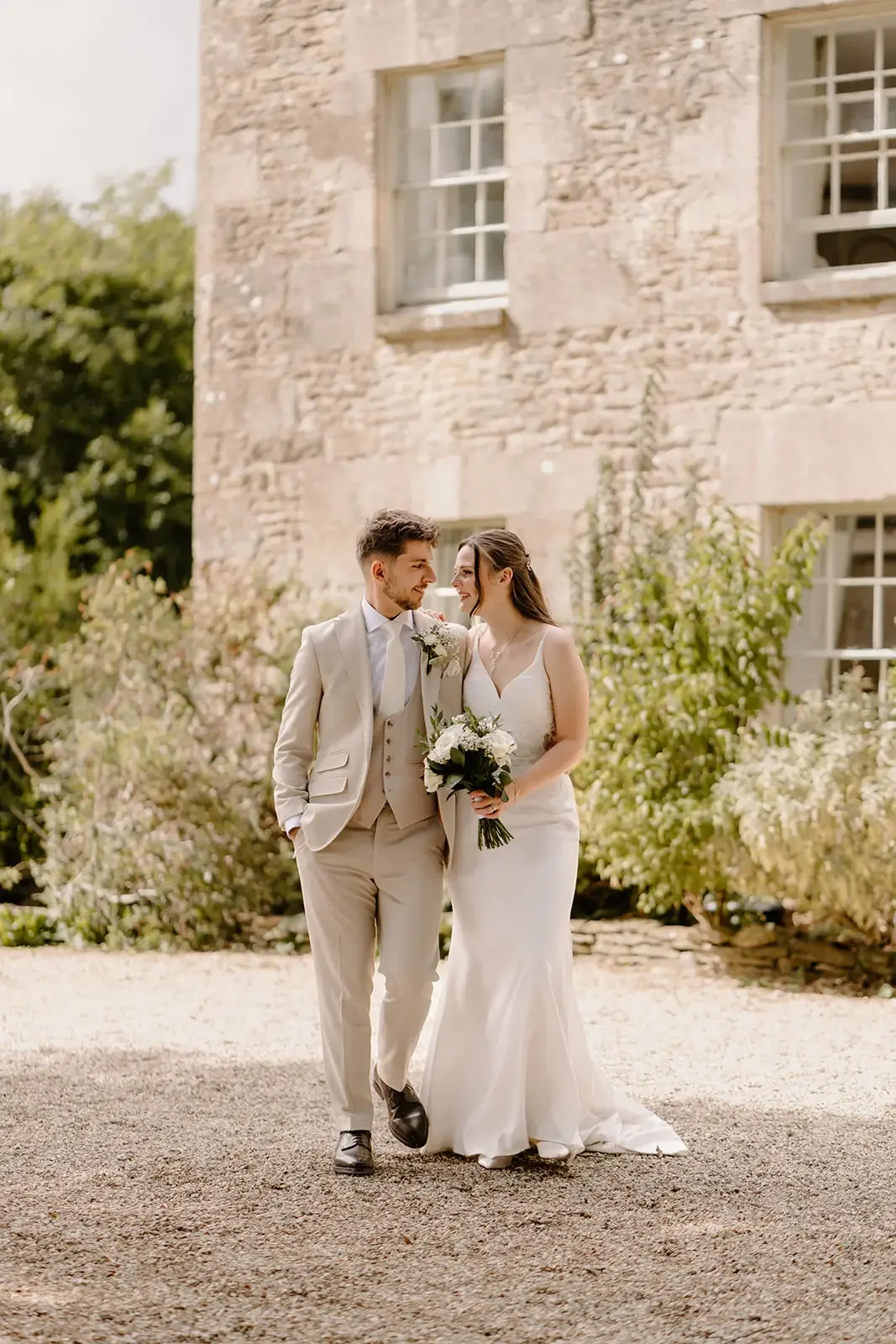 A bride and groom walking together outdoors in a garden with a stone building in the background, the bride holding a bouquet of white flowers.