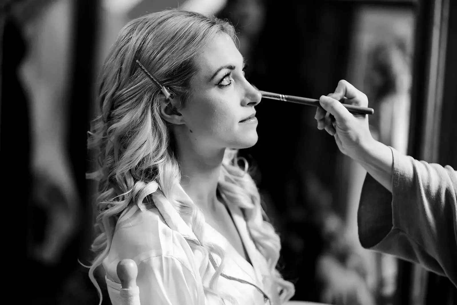 A woman with blonde, curly hair is having her makeup done. Her eyes are looking upward as a makeup artist applies eyeshadow with a brush. The woman is wearing a light-colored jacket, and there are hair clips in her hair. The background is blurred.