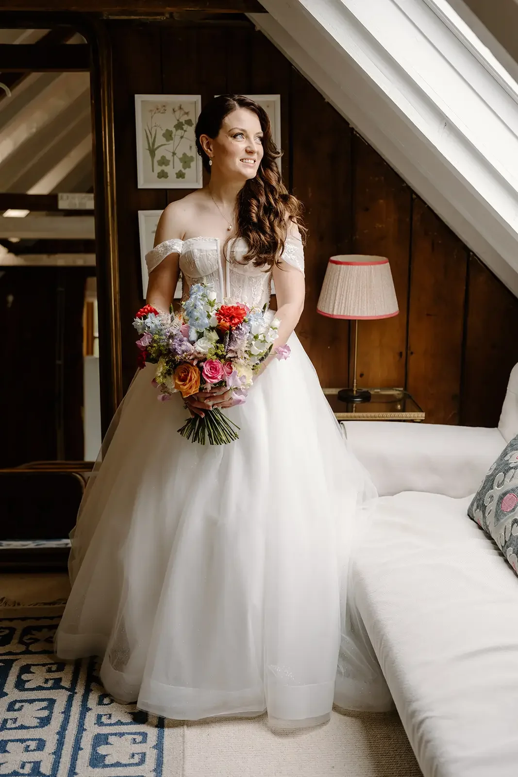 A bride in a white wedding dress holding a colorful bouquet of flowers standing in a cozy room with wooden walls and a sloped ceiling, next to a sofa and a table lamp.