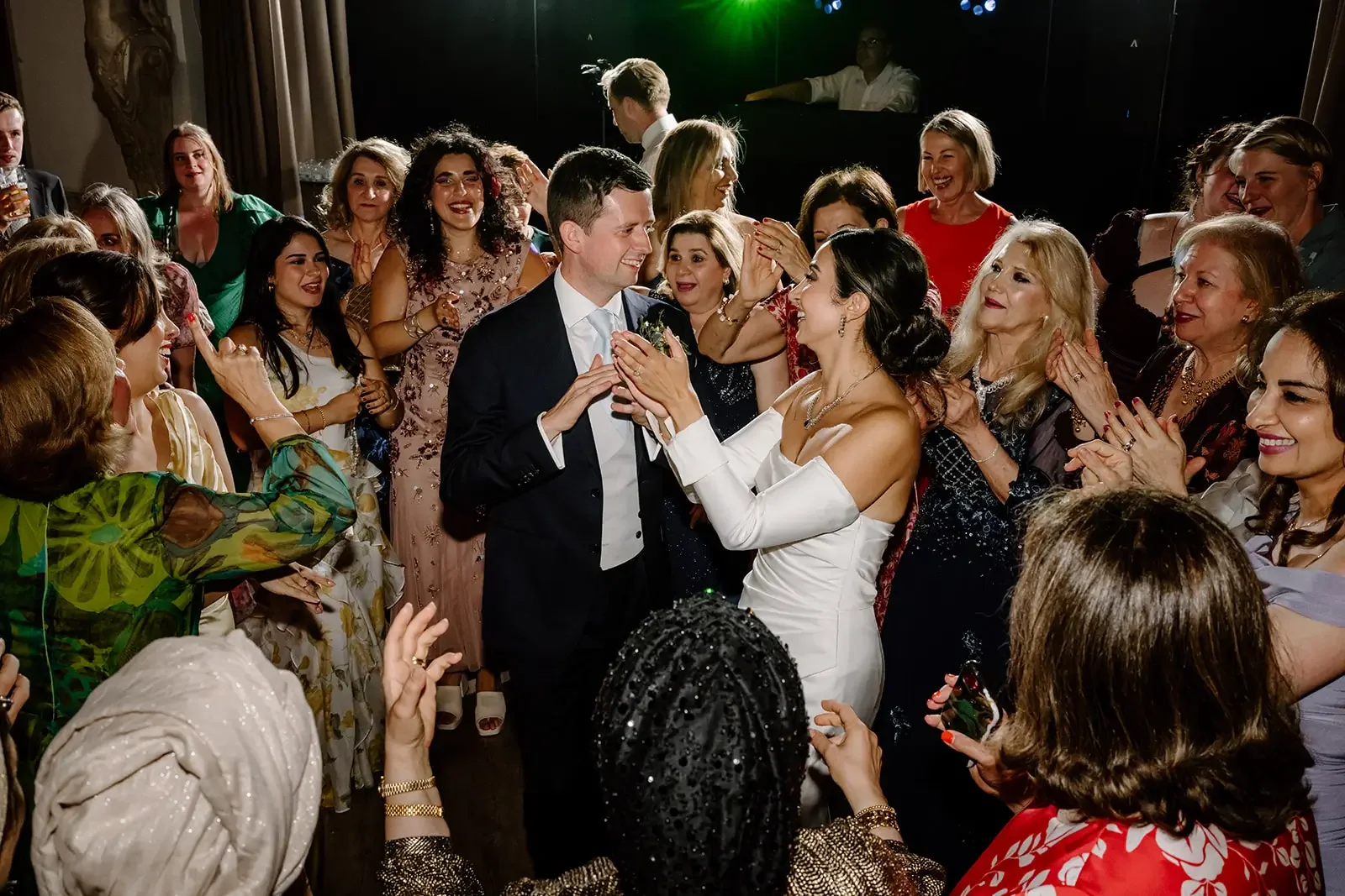 A bride and groom at their wedding reception surrounded by guests, with the bride wearing an off-the-shoulder white gown and the groom in a black tuxedo, celebrating on the dance floor.
