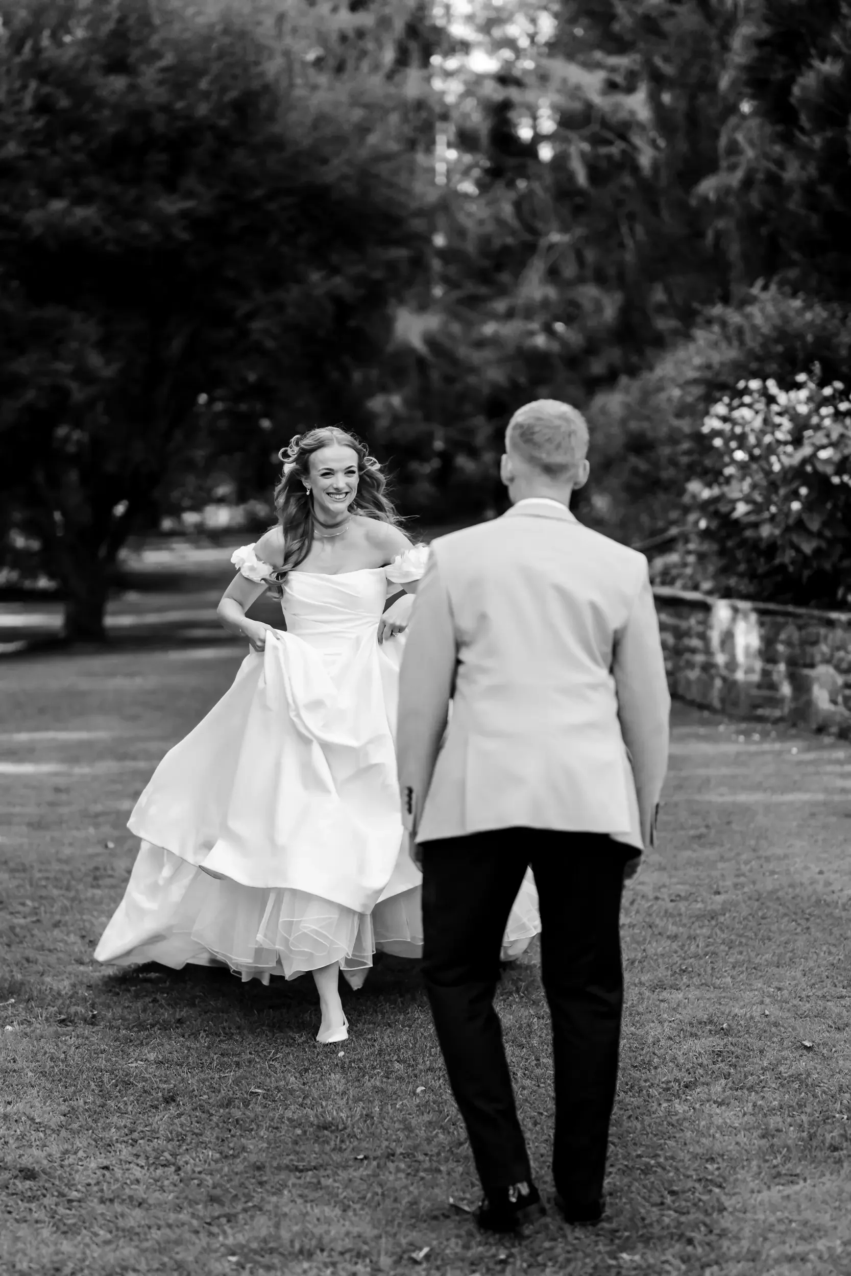 A bride in a wedding dress smiling and walking towards a groom in a suit outdoors.