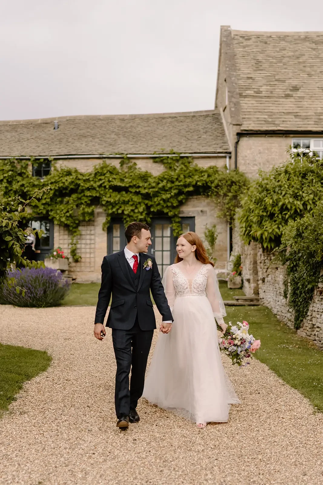 A bride and groom walking hand-in-hand outdoors in a garden courtyard, smiling at each other, with a rustic stone house covered in greenery in the background.