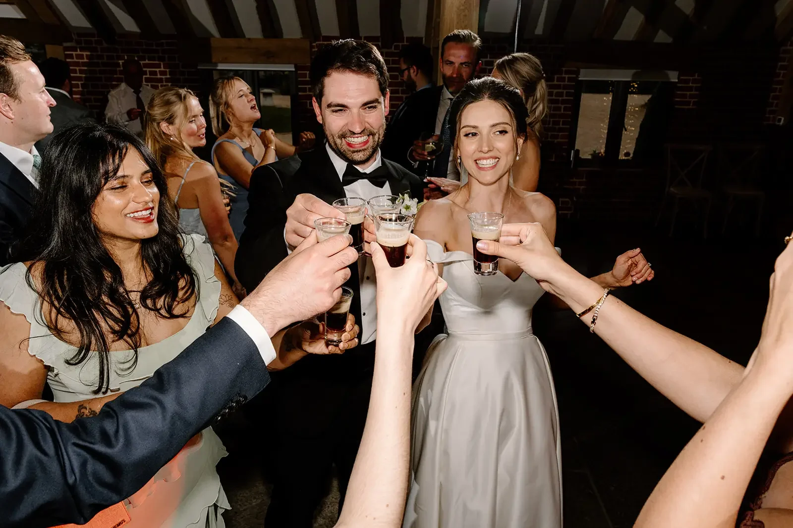 Happy wedding reception with guests toasting drinks, bride in white gown and groom in tuxedo, indoor setting with brick walls.